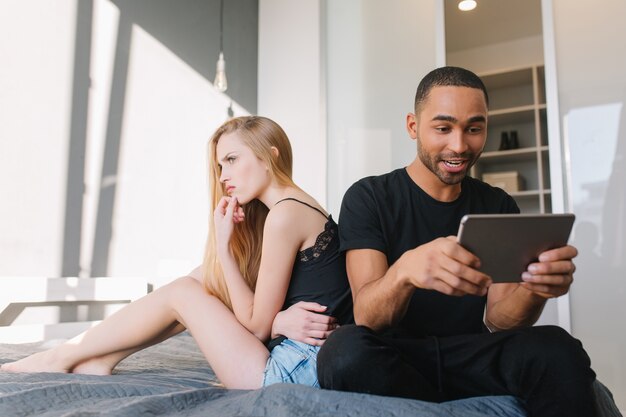Funny lovely moments at home of attractive upset funny young woman looking to window on bed near excited happy guy playing with tablet. Relationship, offended girl, having fun