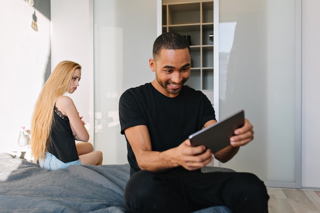 Funny lovely moments at home in modern apartment of attractive upset funny young woman on bed looking at excited happy guy playing with tablet. Relationship, offended girl, having fun