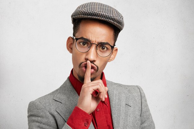 Funny male with dark healthy skin wears round spectacles, keeps finger on lips, makes silence sign