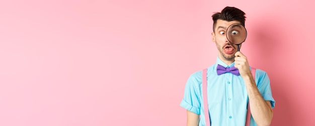 Free photo funny man in bowtie look through magnifying glass squinting and making silly faces standing on pink