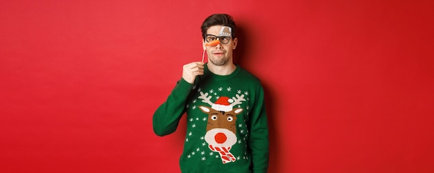 Funny man in christmas sweater and party mask celebrating winter holidays showing funny faces standi...