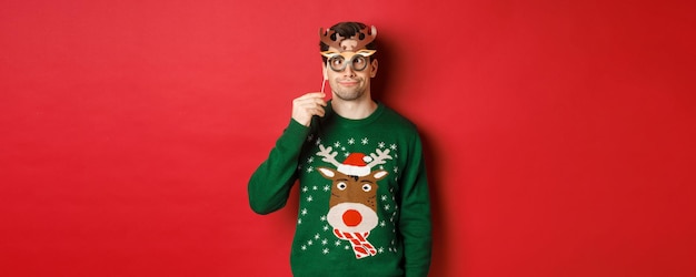 Free photo funny man in christmas sweater and party mask celebrating winter holidays showing funny faces standi...