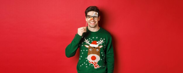 Funny man in christmas sweater and party mask, celebrating winter holidays, smiling happy, standing over red background