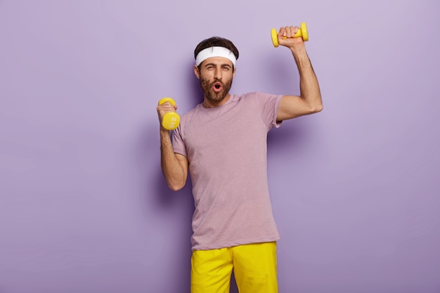 Funny man has fun, exercises with dumbbells, dressed in active wear, motivated for healthy lifestyle, has regular training in morning