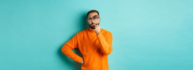 オレンジ色のセーターに立っている何かを検査する虫眼鏡を通して真剣に見える変な男