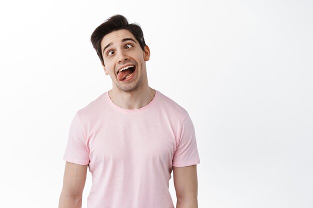 Funny man showing grimaces and tongue, making fun faces playfully, fool around, standing against white wall
