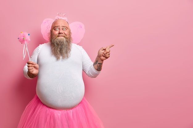 Free photo funny man wears fairy costume, invites you on holiday or costume party, indicates right at blank space, holds magic wand