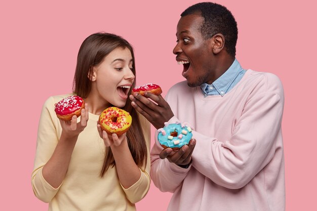 Funny mixed race young woman and man taste delicious donuts, like sweet dessert, bite pastry, stand closely, isolated over pink space