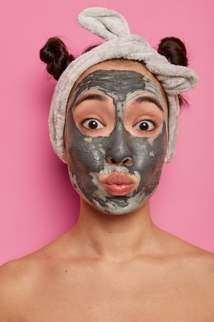 面白い混血の若い女性は唇を吐き出し、顔の粘土マスクを適用し、直接見て、2つのコーマパンを持って、屋内で裸でポーズをとり、新しい美容製品を試し、新鮮なきれいな肌を持ってうれしいです。