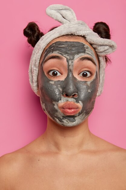 Funny mixed race young woman pouts lips, applies facial clay mask, looks directly , has two combed buns, poses naked indoor, tries new beauty product, glad to have fresh clean skin.