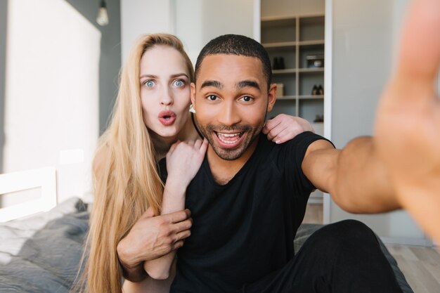 Funny moments of cute couple having fun, making selfie on bed in modern appartment. Young woman with long blonde hair, true emotions, in love, wife, husband, relationship, at home