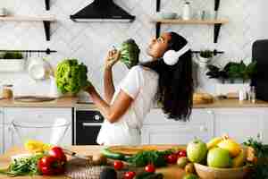 Free photo funny mulatto woman in big wireless headphones is dancing with salad leaves and broccoli on the modern kitchen near table full of vegetables and fruits