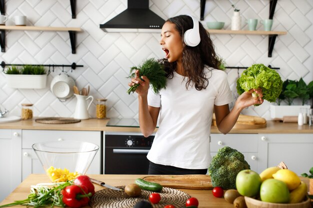 Смешная женщина-мулатка в больших беспроводных наушниках поет на воображаемом зелени микрофона на современной кухне возле стола, полного овощей и фруктов