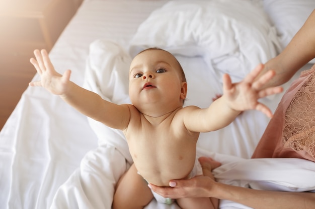 Funny nice little baby woman stretching hands sitting on bed with mom at home.