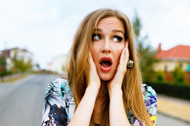 Funny outdoor portrait of crazy hipster blonde woman, put her hands to her cheeks, surprised emotions.