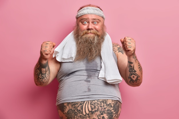 Free photo funny overweight man being sweaty after intensive cardio, raises clenched fists, dressed in sportswear, does morning exercises to loose weight puts all efforts to be fit and healthy. sport and obesity