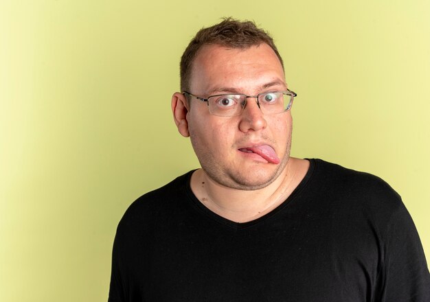 Funny overweight man in glasses wearing black t-shirt sticking out tongue over light