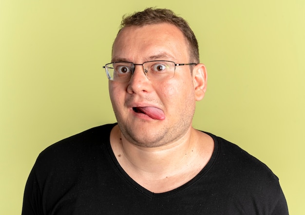 Funny overweight man in glasses wearing black t-shirt sticking out tongue over light