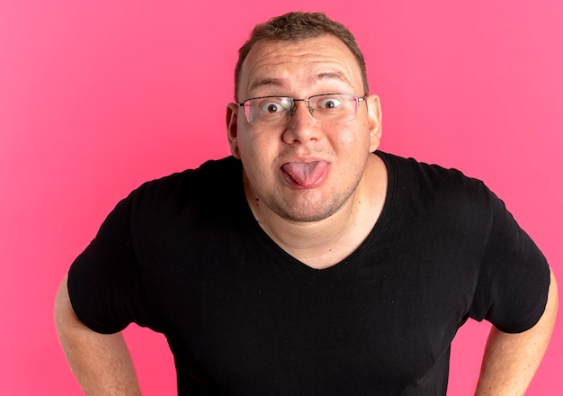 Funny overweight man in glasses wearing black t-shirt  sticking out tongue standing over pink wall