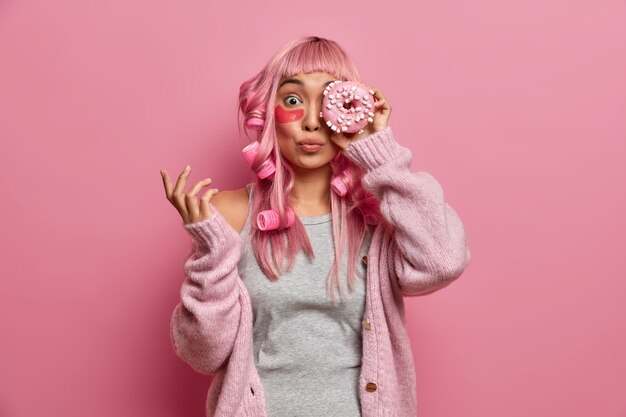 Funny pink haired Asian woman has rollers on head, covers eye with tasty sweet doughnut, wears collagen patches to reduce wrinkles