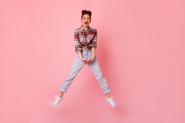 Ragazza divertente del pinup in blue jeans che salta e che guarda l'obbiettivo. vista integrale della giovane donna in camicia a scacchi che balla sullo spazio rosa.