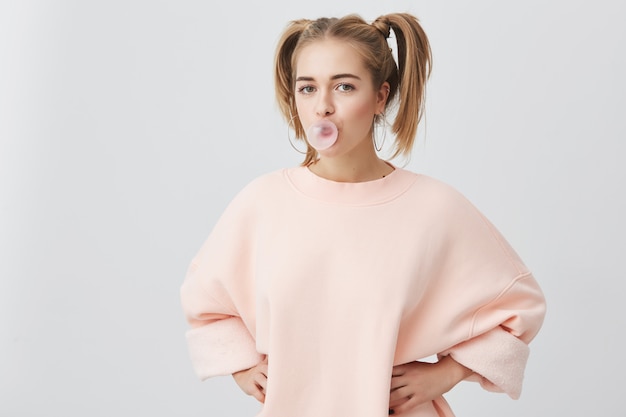 Free photo funny playful fair-haired female teenager with two ponytails wearing pink long-sleeved sweater having joyful expression, with chewing gum bubble in her mouth, isolated