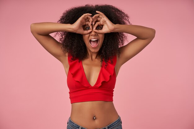 Funny portrait of pretty young dark skinned woman with casual hairstyle wearing red top and jeans, fooling on pink and folding eyewear from raised hands, smiling broadly