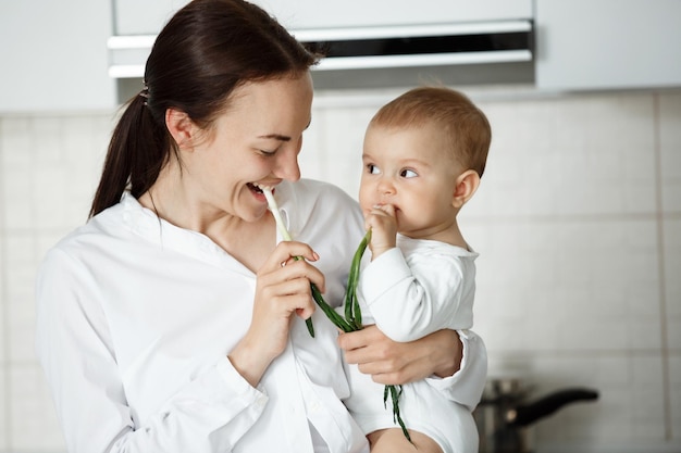 Funny portrait of young beautiful mother looking at her little boy while he looking at her with curious expression They eating green onion at kitchen