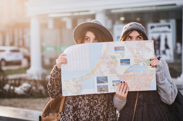 Funny positive image of fashionable women on sunny street having fun in city, hiding behind the citymap. Travelling together, best friends, get a lost in big city, true emotions.