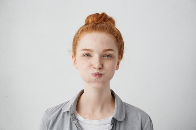 Funny pretty girl puffing out her cheeks against white wall. Headshot of charming red haired young woman making mouths while having fun indoors. People, lifestyle, youth and happiness