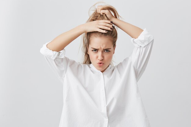 Funny pretty girl with frowned face and darkeyes, tying her blonde hair in ponytail, getting ready before going out to cinema with friends. Young Caucasian female model posing