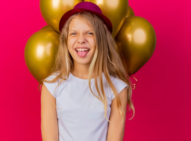 Смешная симпатичная маленькая девочка в праздничной шляпе с кучей воздушных шаров счастливой и взволнованной