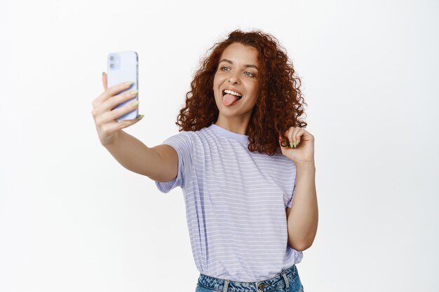 Funny redhead girl making selfie on mobile phone, photo filter on smartphone app, showing tongue on white.