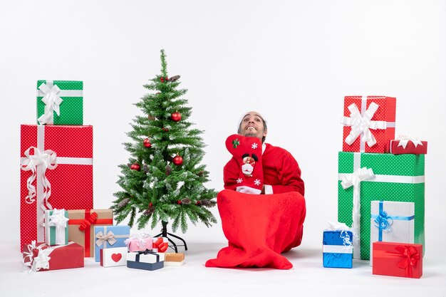 Free photo funny santa claus sitting on the ground and wear christmas sock near gifts and decorated new year tree on white background stock photo