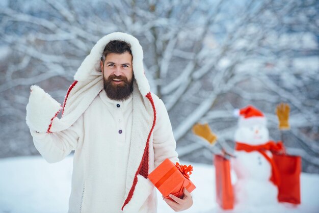 冬の天気で赤いギフトボックスでポーズをとって面白いサンタの男。うれしそうな父は、ウィンターパークで贈り物と雪だるまを楽しんでいます。