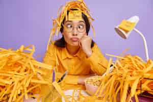 Free photo funny schoolgirl prepares for test at home has sleepless night works late hours makes grimace has busy working schedule surrounded by paper wastes isolated over purple wall