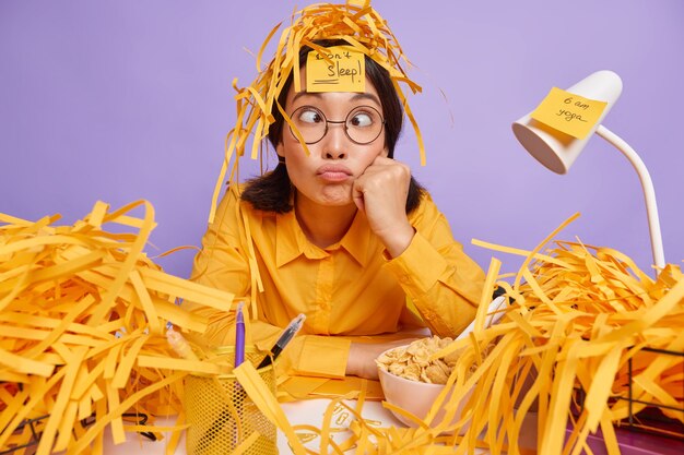 Funny schoolgirl prepares for test at home has sleepless night works late hours makes grimace has busy working schedule surrounded by paper wastes isolated over purple wall