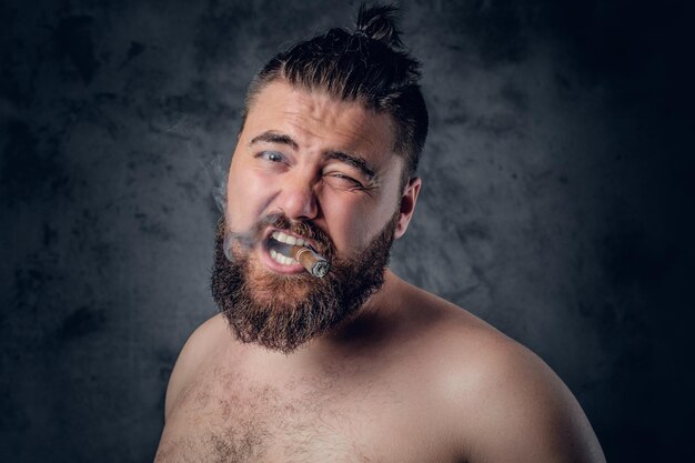 Funny shirtless bearded male smoking a cigar on grey background.