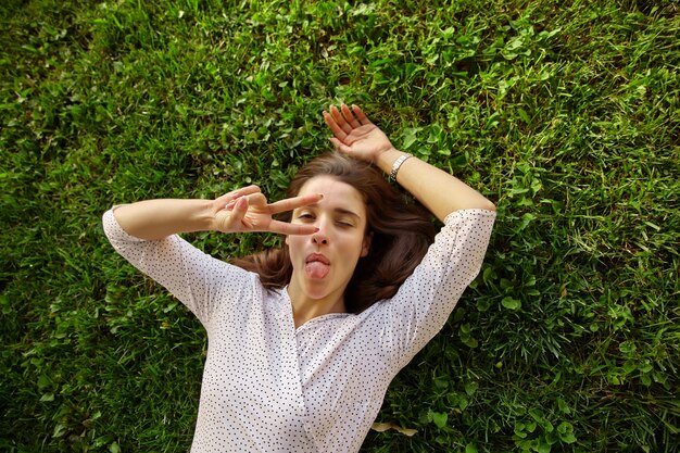 Funny shot of young dark haired pretty woman posing outdoor in white polka-dot clothes, showing tongue and raising hand with victory gesture while looking