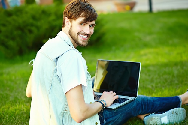 Смешной улыбающийся битник красавец парень в стильной летней ткани в траве с ноутбуком