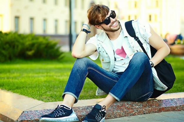 公園の芝生の上に座って通りにスタイリッシュな夏布で面白い笑顔ヒップスターハンサムな男男