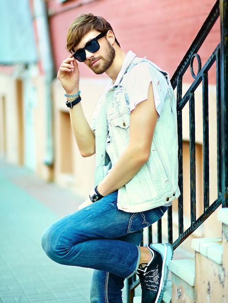 Funny smiling hipster handsome man guy in stylish summer clothes in the street posing near colorful bright wall in sunglasses