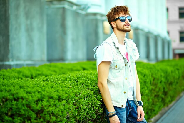 Funny smiling hipster handsome man guy in stylish summer clothes in the street posing near green city 