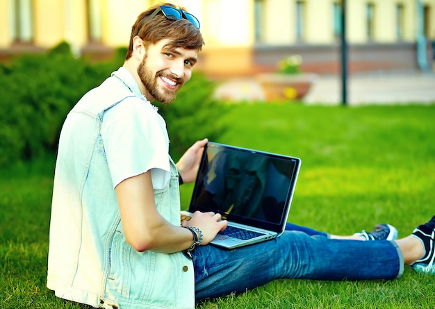 Funny smiling hipster handsome man guy in stylish summer clothes in the street posing sitting on grass with notebook