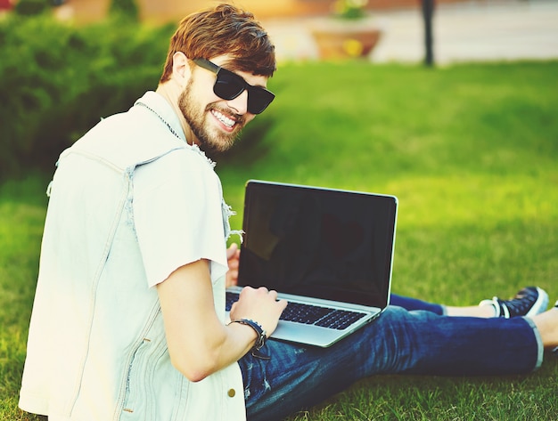 Смешной улыбающийся битник красавец парень в стильной летней одежде на улице сидит на траве в парке с ноутбуком
