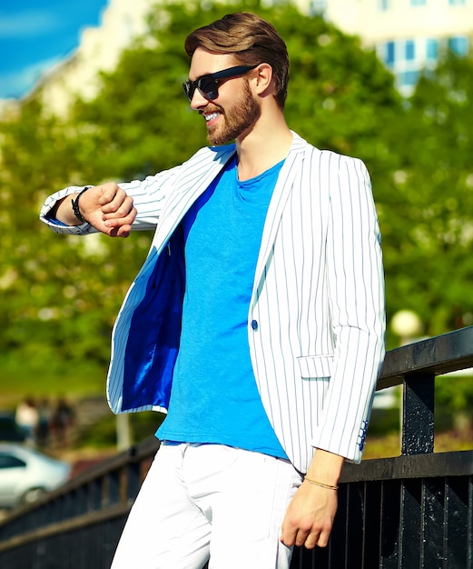 Funny smiling hipster handsome man in stylish summer white suit posing on street in sunglasses, looking at watches