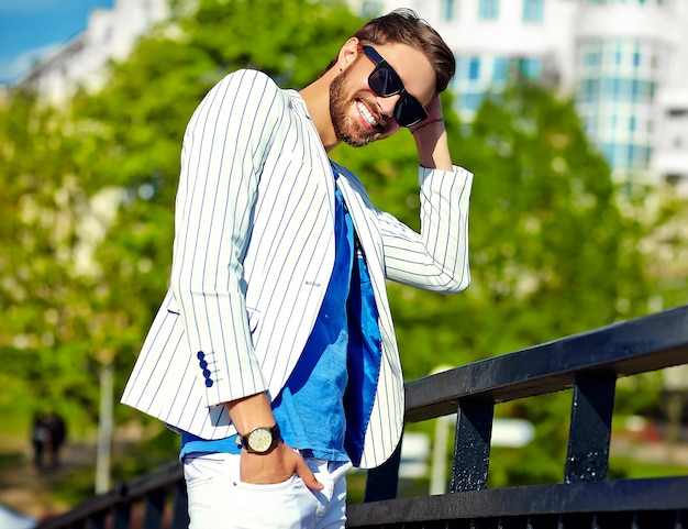 Uomo bello sorridente divertente dei pantaloni a vita bassa in vestito bianco di estate alla moda che posa in via in occhiali da sole