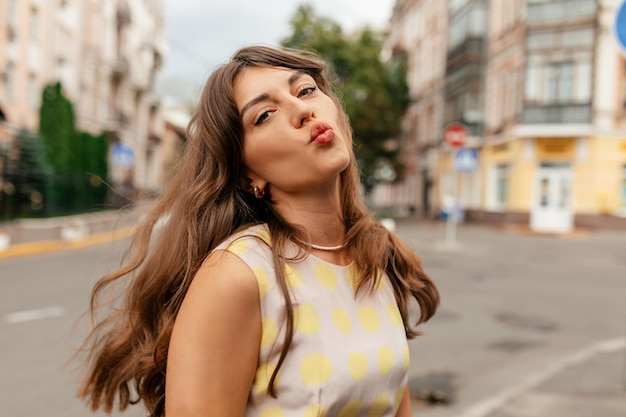 Funny spectacular lovely girl with wavy flying hair from wind walking in the city and sending a kiss on blurred city background European young girl posing tenderly