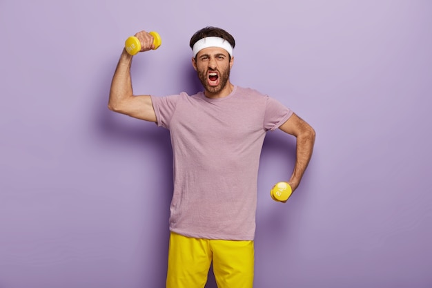 面白いスポーツマンは、ダンベルで腕を上げ、感情的に叫び、強くてスポーティーな気分になり、紫色のTシャツと黄色のショートパンツを着て、屋内に立っています。男はジムで運動し、運動をします。ボディービル