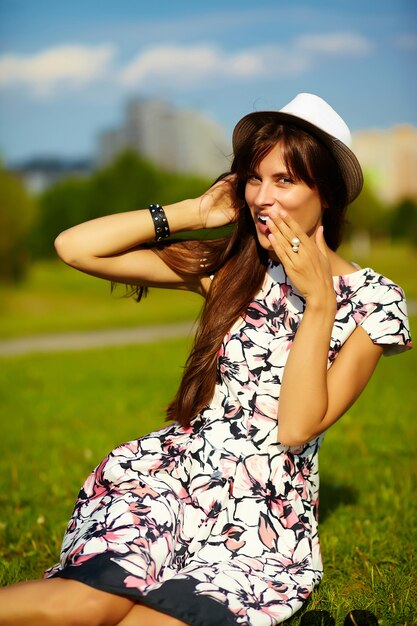 Смешная стильная сексуальная улыбающаяся красивая модель молодой женщины в ярком летнем платье из ткани битника в шляпе сидит на траве в парке